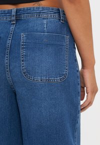 Denim jeans in a medium blue wash, featuring a single back pocket, belt loops, and a smooth texture with subtle stitching details.
