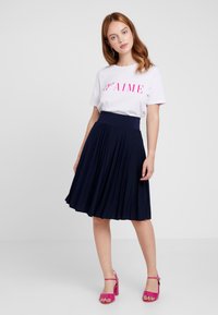 Weißes T-Shirt mit pinkem Druck, kombiniert mit einem navyblauen Faltenrock. Das Modell trägt rosa Absatzsandalen. Einfache Hintergrund.