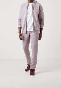 Sudadera con cremallera color púrpura claro con capucha y logo, combinada con pantalones a juego y zapatillas deportivas burdeos con suelas verdes. Textura suave.