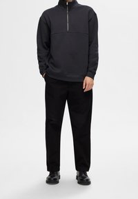 Mörkgrå zip-up sweatshirt med hög krage, har en söm på frampanelen; parat med lösa svarta byxor och svarta skor.