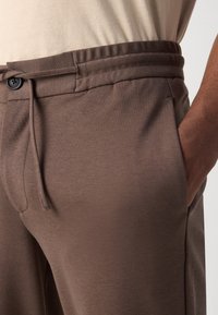 Brune bomull joggebukser med justerbar snor i livet, flat front og sidelomme. Stoffet har en glatt tekstur og matt finish.