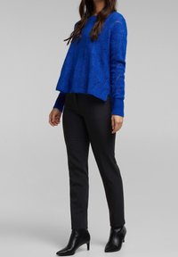 Blauer gestrickter Pullover mit strukturiertem Muster, lockerem Sitz, gerippten Bündchen; kombiniert mit schwarzen Slim-Fit-Hosen und schwarzen Ankle-Boots mit Absatz.