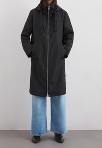 Svart lång jacka med dragkedja fram, två sidofickor och avtagbar huva. Kombinerad med ljusblå vida jeans och svarta ankelkängor.