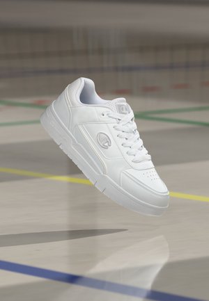 Weißer Ledersneaker mit Schnürsenkeln und Markenlogo, der über einem polierten Hallensportboden mit farbigen Linien schwebt.