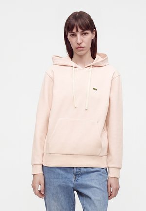Jeune femme aux cheveux bruns raides portant un sweat à capuche rose pâle et un jean bleu sur un fond blanc uni.