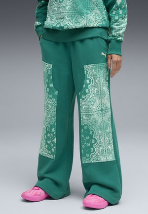 Persoon in groene wijde broek met paisley-patroon patches en bijpassende sweatshirt, gecombineerd met felroze instapschoenen.