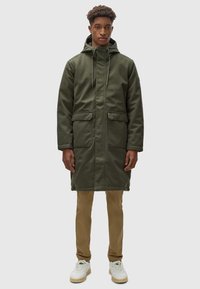 Parka verde oliva con capucha, dos bolsillos frontales, cierre de cremallera y cordones. Lleva pantalones beige y zapatillas blancas. Textura suave.