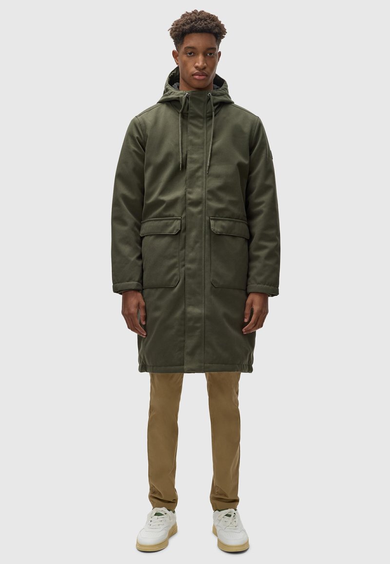Parka verde oliva con capucha, dos bolsillos frontales, cierre de cremallera y cordones. Lleva pantalones beige y zapatillas blancas. Textura suave.