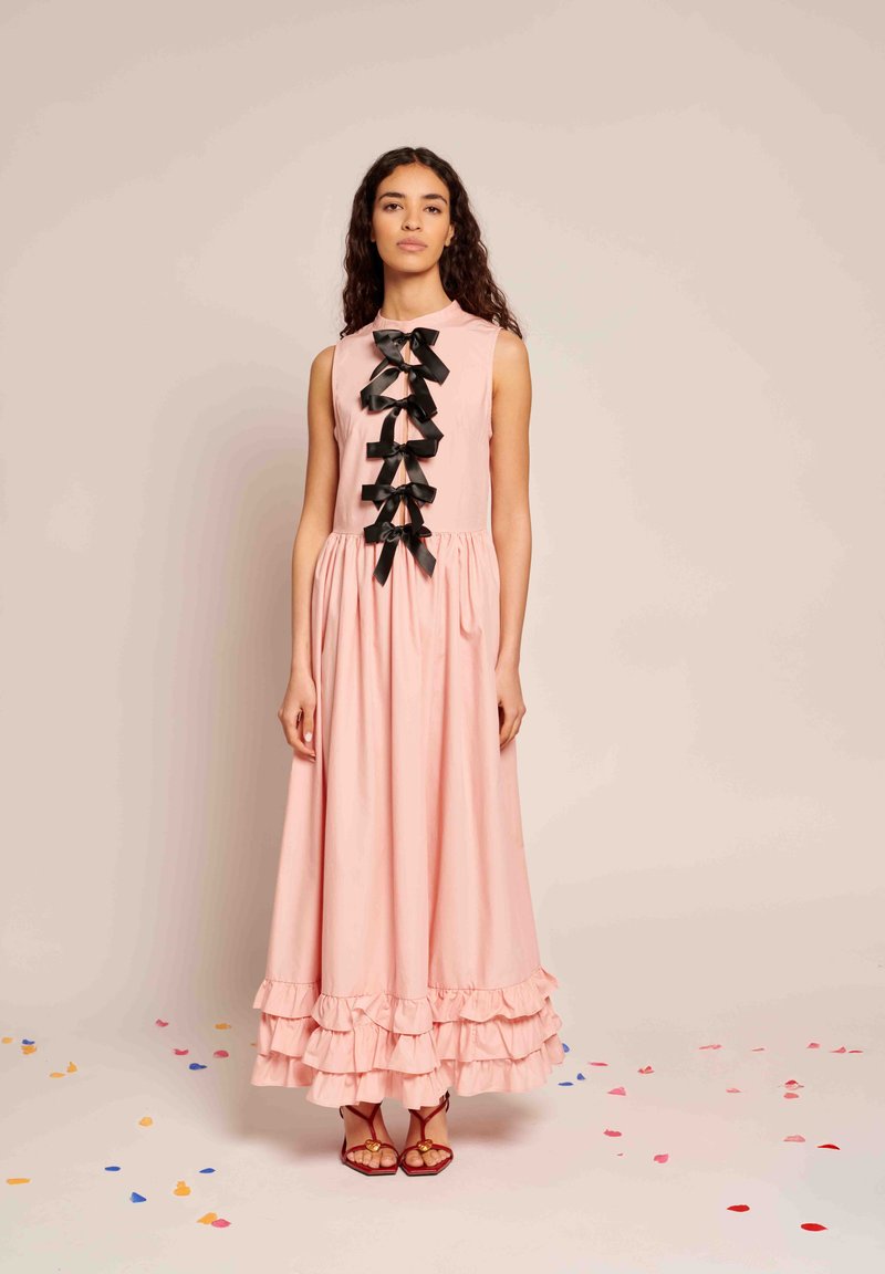 Hosszú rózsaszín ruha, formázott bodice-szal, kerek nyakkivágással és fekete szatén masnikkal az elején. Rózsa fodros szegéllyel és puha szövet textúrával.