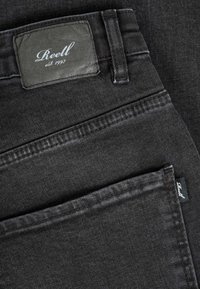 Close-up of black denim jeans showing a leather brand patch with "Reell est. 1997" and a small fabric label on the back pocket.