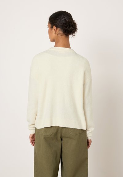 Pull en maille crème avec une coupe décontractée et des accents côtelés, associé à un pantalon large vert olive. Texture douce, longueur légèrement raccourcie.
