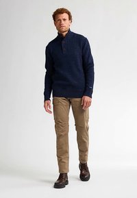 Maglione lavorato a maglia blu navy con collo alto e tre bottoni, abbinato a pantaloni cargo beige e stivali stringati marrone scuro.
