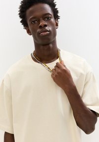 Tricou oversized bej, fabricat dintr-un material moale, cu mâneci scurte. Modelul poartă lanțuri suprapuse, unul auriu și unul argintiu.