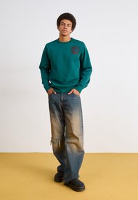 Ung mann står med hendene i lommene iført en teal hettegenser, vide falmede jeans med et knesår, og svarte støvler på gulvet.