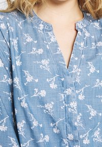 Jasnoniebieska bluzka uszyta z teksturowanej tkaniny, zdobiona białym kwiatowym wzorem, z częściowym zapięciem na guziki i okrągłym dekoltem.