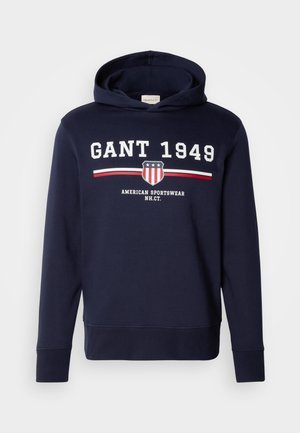Sudadera azul marino con un gráfico frontal que presenta "GANT 1949" y un logo de escudo. Confeccionada en material suave, tiene mangas largas y capucha ajustable con cordón.