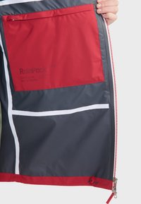 De�štník RainPack má šedou vnější stranu, červenou vnitřní kapsu, bílé detaily a zip s červenými prvky. Materiál je voděodolný.