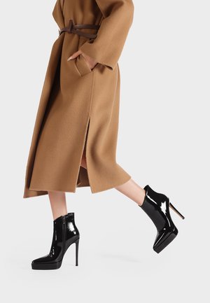 Stivaletti neri in pelle lucida con platfom, tacco a stiletto e cerniera laterale; indossati con un cappotto beige con cintura. Texture liscia, design elegante.