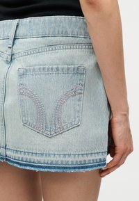 Φούστα μίνι από ανοιχτό μπλε τζιν με ξεθωριασμένο τελείωμα και ραμμένη πίσω τσέπη, φορεμένη με μαύρο τοπ, που δείχνει μερικώς το δεξί χέρι και μπράτσο.