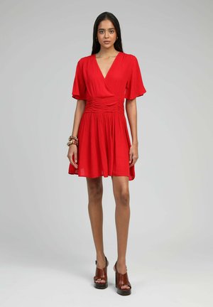 Robe rouge avec un décolleté en V et des manches courtes à volants, présentant une taille froncée et une jupe évasée. Le mannequin porte des sandales compensées marron.