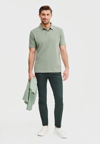 Jasnozielona koszulka polo w połączeniu z ciemnozielonymi spodniami. Osoba trzyma jasnozieloną kurtkę i nosi białe trampki. Prosty design, bez wzorów.