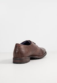 Chaussure habillée pour homme en cuir marron avec lacets à l’avant et petit talon empilé, inclinée pour montrer l’arrière et le côté sur un fond clair uni.