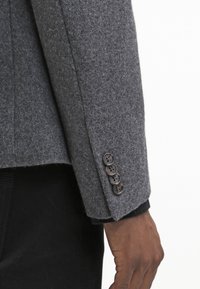 Šedý vlněný blazer s rukávem, na kterém jsou čtyři hnědé knoflíky na manžetě. Textura vypadá jemně a design se vyznačuje přesným střihem.