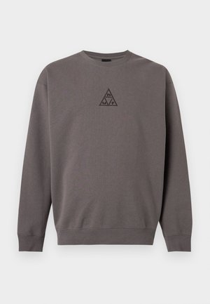 Ung mand iført en grå sweatshirt med et lille trekantet logo, lagdelt over en ternet skjorte, stående mod en ensfarvet baggrund.