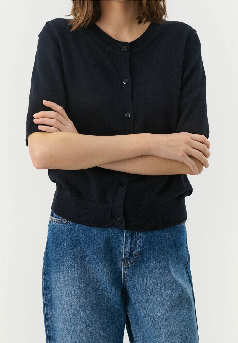Personne portant un cardigan noir à manches courtes boutonné et un jean bleu, bras croisés à la taille devant un fond uni.