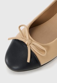 Beige ballerinatossa, jossa on musta pyöreä kärki, ja edessä pieni rusetti. Tasapintainen materiaali, jossa on tikattuja yksityiskohtia reunoilla.