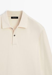 Beżowa koszulka polo z miękkiej bawełny, z klasycznym kołnierzykiem, dwukolorową plisą na guziki oraz pojedynczym czarnym guzikiem jako akcentem.
