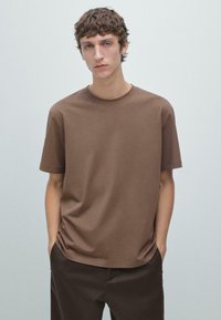 T-shirt marrone a girocollo realizzata in morbido cotone, con maniche corte e vestibilità comoda. Il modello posa con le mani nelle tasche.