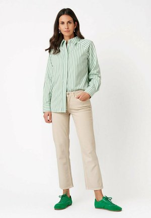 Woman standing with hand in pocket wearing green striped long-sleeve shirt, beige pants, and green sneakers against a plain background.