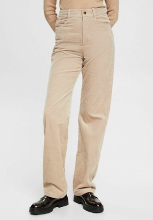 Pantalons en velours côtelé beige taille haute portés avec des mocassins noirs épais et un haut à manches longues beige à côtes, les mains derrière le dos.