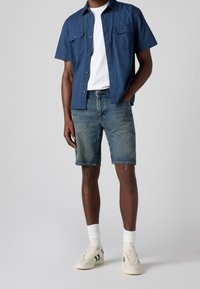 Un homme portant une chemise à manches courtes bleu, un t-shirt blanc, un short en jean, des chaussettes blanches et des baskets blanches avec des rayures vertes.
