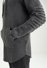 Šedá mikina s dlouhým rukávem a klokánkovou kapsou. Má strukturované, skládací rukávy a kapuci se šňůrkou. Měkký materiál připomínající svetr.