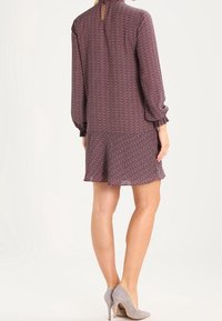 Kortärmad klänning i mörk burgund med ett geometriskt mönster, volangärmar och en nyckelhålsdetalj bak, kombinerad med grå höga klackar.