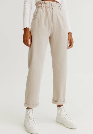 Person wearing beige high-waist paperbag pants with rolled cuffs and white lace-up platform sneakers against a plain background.