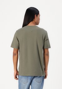 T-shirt in cotone verde oliva con vestibilità rilassata, maniche corte, dettaglio della cucitura sul retro del collo, abbinato a jeans in denim azzurro chiaro.