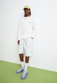Witte lange mouwen blouse met een zak, lichtblauwe shorts, grijze en paarse sneakers, en een groene pet. Eenvoudige, casual streetwear look.