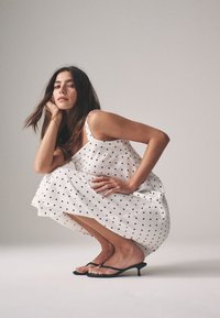 White polka dot dress with thin straps, knee-length, lightweight fabric. Black heeled sandals. Model squatting, with long dark hair.