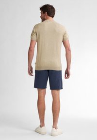 Beige gestricktes kurzärmliges Hemd mit gerippten Bündchen, kombiniert mit navy-blauen gestreiften Shorts. Das Model steht mit dem Rücken zur Kamera und zeigt eine lässige Passform.