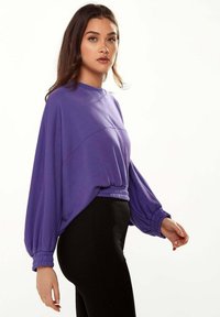 Purple sweatshirt with a rounded neckline, dropped shoulders, and elasticated hem. Features long, billowing sleeves and a relaxed fit.