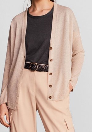 Frau trägt eine beige Knopfleiste Cardigan über einem schwarzen Oberteil, beige Hosen mit Cargotaschen und einen schwarzen gewebten Gürtel mit einer runden Schnalle.