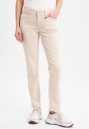 Personne portant un pantalon beige droit, la main dans la poche, et des baskets blanches avec des semelles bordeaux sur fond blanc.