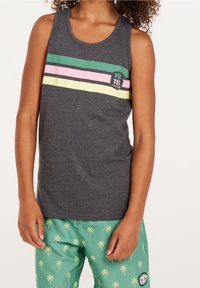 Persona che indossa una maglietta senza maniche grigio scuro con strisce verdi, rosa e gialle e pantaloni corti verdi con motivi di palme gialle.