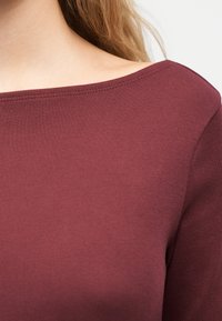 Γυναίκα που φοράει μπορντό μπλούζα με λαιμόκοψη σκάφος, δείχνοντας το λαιμό, τον ώμο και μέρος από τα μακριά ξανθά μαλλιά της, με φόντο λευκό.