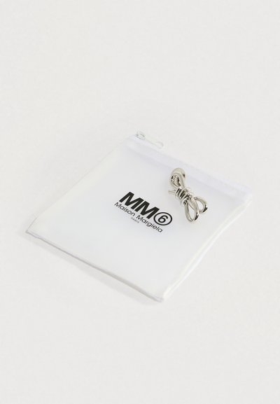 Ezüst, csavart fém gyűrű, fehér tasakon bemutatva, amelyen fekete MM6 Maison Margiela logó látható, egyszínű fehér háttéren.