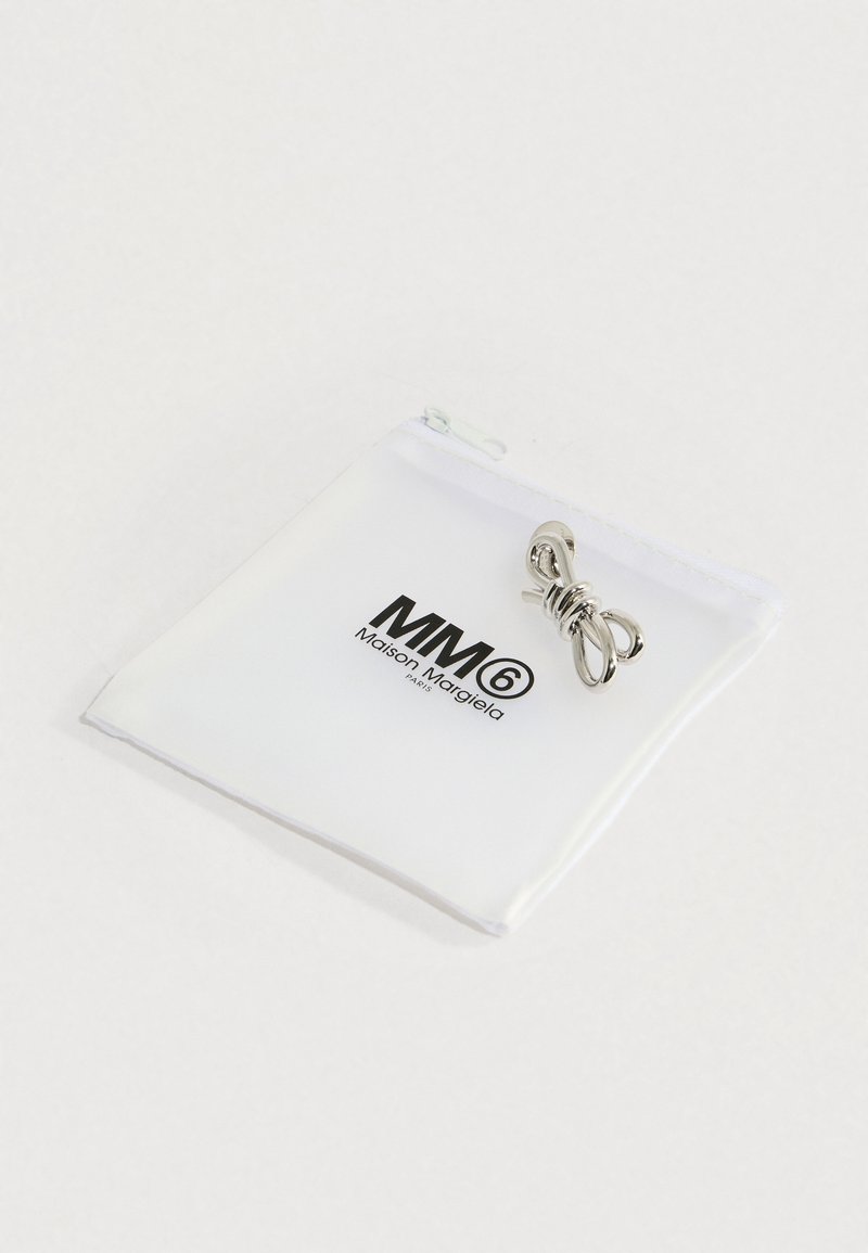 Anillo de metal plateado retorcido exhibido sobre una bolsa blanca con el logo negro de MM6 Maison Margiela, sobre un fondo blanco liso.
