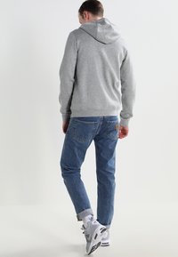 Muž v šedé mikině s kapucí, modrých džínách a bílých teniskách odchází proti jednoduchému bílému pozadí.